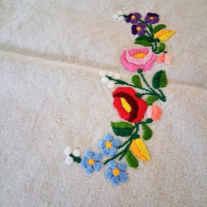 May include: Close-up of a beige linen fabric with embroidered floral design. The embroidery features a variety of colorful flowers, including red, pink, purple, and blue, with green leaves and yellow accents. The flowers are arranged in a curved pattern.