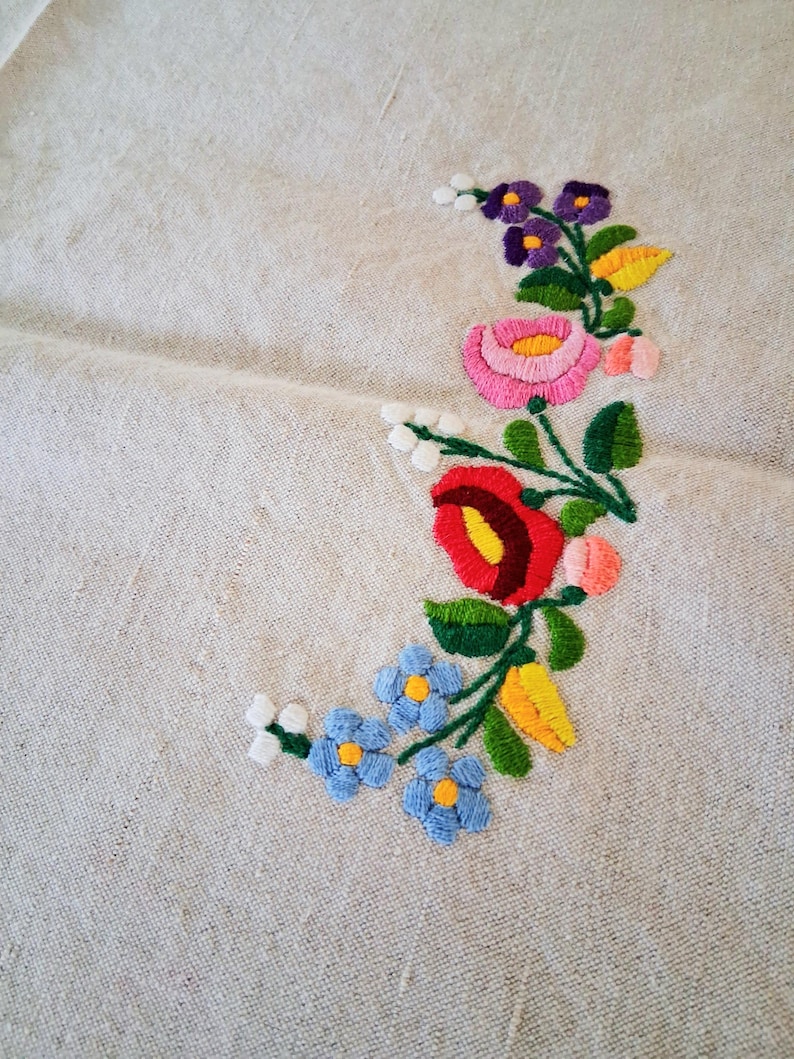 May include: Close-up of a beige linen fabric with embroidered floral design. The embroidery features a variety of colorful flowers, including red, pink, purple, and blue, with green leaves and yellow accents. The flowers are arranged in a curved pattern.