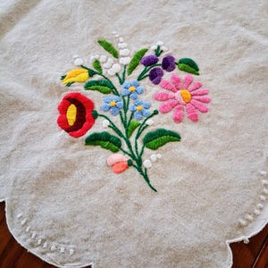 May include: Embroidered floral design on a beige linen tablecloth. The colorful bouquet features red, yellow, blue, pink, and purple flowers with green leaves. The tablecloth has a scalloped edge with white stitching.