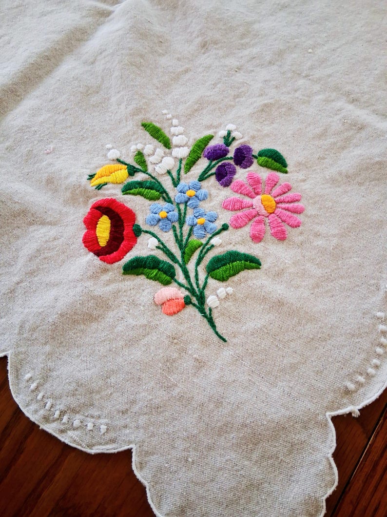 May include: Embroidered floral design on a beige linen tablecloth. The colorful bouquet features red, yellow, blue, pink, and purple flowers with green leaves. The tablecloth has a scalloped edge with white stitching.