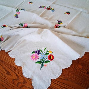 May include: Cream-colored embroidered tablecloth with a scalloped edge. The tablecloth features colorful floral embroidery in pink, purple, blue, and red. The cloth is draped over a wooden table.