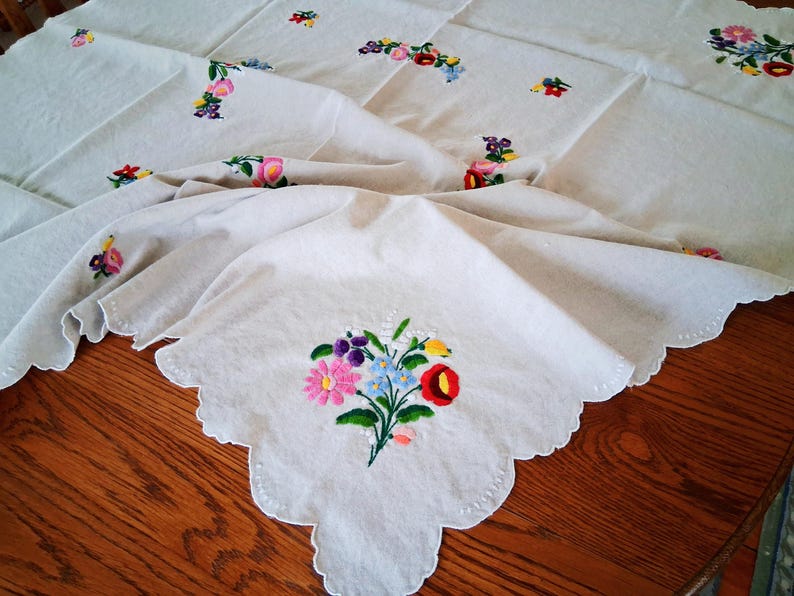 May include: Cream-colored embroidered tablecloth with a scalloped edge. The tablecloth features colorful floral embroidery in pink, purple, blue, and red. The cloth is draped over a wooden table.