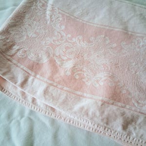 May include: A folded pink and white damask tablecloth. The tablecloth features a central pink panel with a white floral design. The edges are a lighter pink with a simple border. The fabric appears soft and slightly textured.