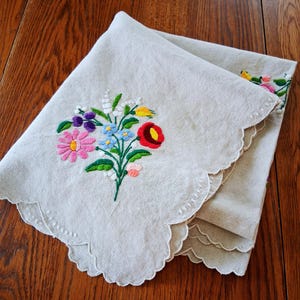 May include: A folded, off-white linen cloth with a scalloped edge. The cloth is embroidered with a colorful floral bouquet featuring pink, red, blue, and yellow flowers. The cloth is folded on a wooden surface.