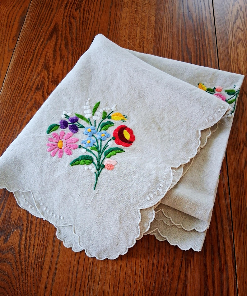 May include: A folded, off-white linen cloth with a scalloped edge. The cloth is embroidered with a colorful floral bouquet featuring pink, red, blue, and yellow flowers. The cloth is folded on a wooden surface.