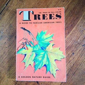 May include: A softcover book titled Trees: A Guide to Familiar American Trees, A Golden Nature Guide. The orange cover features a large illustration of green and yellow maple leaves with brown seeds hanging from a branch, with black text.