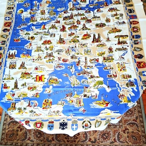 May include: A vintage map-themed tablecloth featuring a detailed illustration of Europe. The map is in blue and beige, with colorful drawings of landmarks and flags around the border. The tablecloth is rectangular and made of fabric.