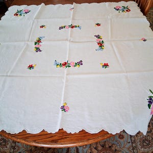 May include: White tablecloth with embroidered floral designs in pink, purple, yellow, and blue. The scalloped edge adds a decorative touch. The tablecloth is spread out on a wooden table, showcasing the detailed needlework.
