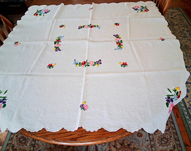 May include: White tablecloth with embroidered floral designs in pink, purple, yellow, and blue. The scalloped edge adds a decorative touch. The tablecloth is spread out on a wooden table, showcasing the detailed needlework.