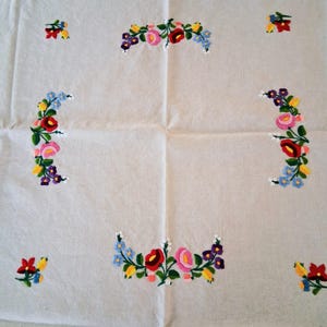 May include: A white tablecloth with embroidered floral designs. The embroidery features red, pink, yellow, blue, and purple flowers arranged in curved patterns and small clusters across the fabric. The cloth is folded.