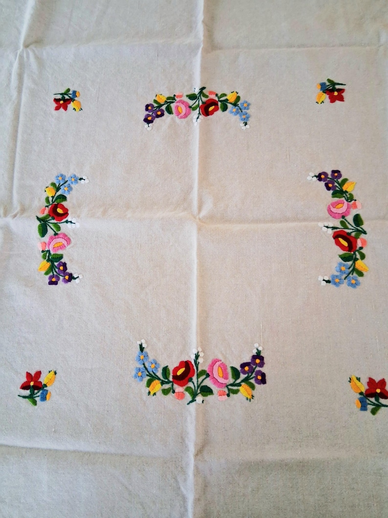 May include: A white tablecloth with embroidered floral designs. The embroidery features red, pink, yellow, blue, and purple flowers arranged in curved patterns and small clusters across the fabric. The cloth is folded.