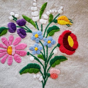 May include: Close-up of a floral embroidery design featuring a bouquet of colorful flowers. The design includes pink daisies, purple tulips, blue forget-me-nots, a red poppy, and white lily of the valley. The embroidery is on a beige linen fabric.