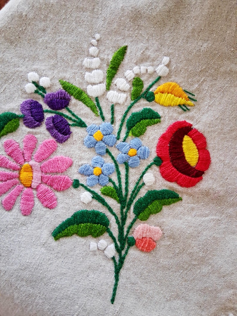 May include: Close-up of a floral embroidery design featuring a bouquet of colorful flowers. The design includes pink daisies, purple tulips, blue forget-me-nots, a red poppy, and white lily of the valley. The embroidery is on a beige linen fabric.