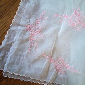 May include: A sheer, white fabric square with a scalloped edge. Pink floral embroidery decorates the corners and center, creating a delicate, vintage aesthetic. The fabric appears lightweight and airy, suitable for decorative purposes.