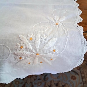May include: White linen table runner with embroidered floral design. The embroidery features white flowers with yellow centers and delicate leaves. The edge is scalloped and finished with a crocheted trim, adding a touch of elegance to the piece.