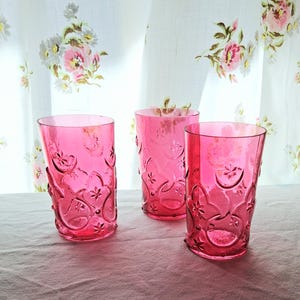 May include: Three pink drinking glasses with embossed floral patterns. The glasses are made of a translucent material and are arranged on a white surface. The background features a floral patterned curtain.