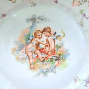 May include: A white porcelain plate featuring a central illustration of two cherubs sitting together in a garden setting. The plate has a fluted rim decorated with small purple floral sprigs and gold lattice patterns along the outer edge.