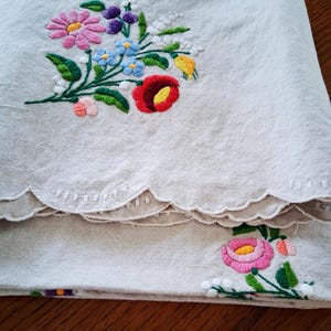 May include: Beige linen fabric with colorful floral embroidery. The embroidery features a bouquet of pink, blue, red, yellow, and purple flowers with green leaves. The edge of the fabric has a scalloped design.