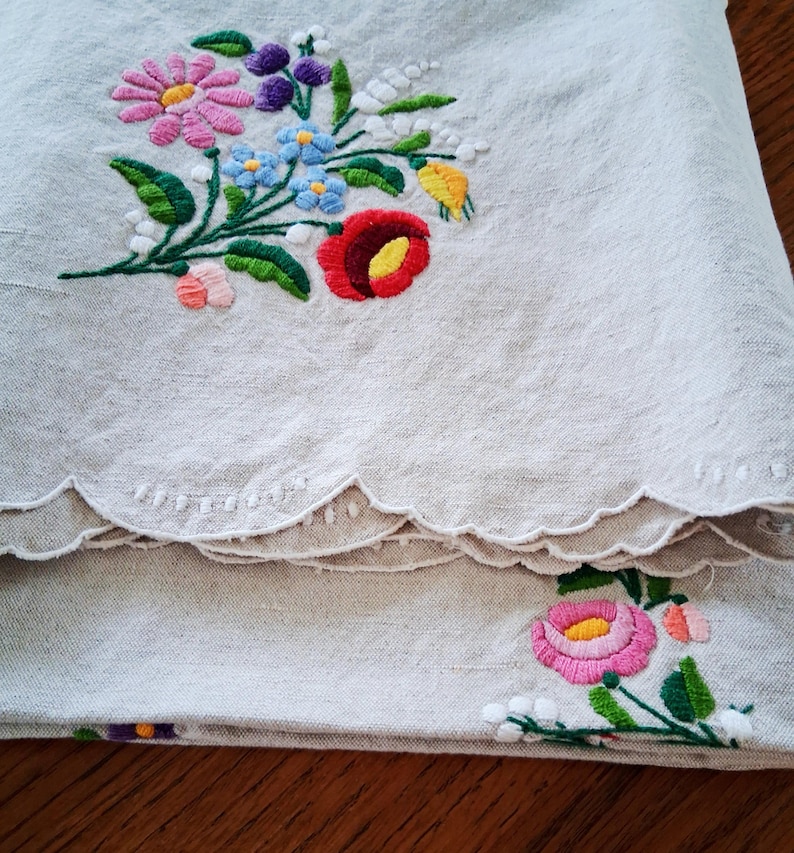 May include: Beige linen fabric with colorful floral embroidery. The embroidery features a bouquet of pink, blue, red, yellow, and purple flowers with green leaves. The edge of the fabric has a scalloped design.