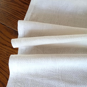 May include: Close-up of a folded white linen table runner. The fabric has a subtle floral pattern and a textured weave. The runner is draped over a wooden surface, showcasing its folds and texture.