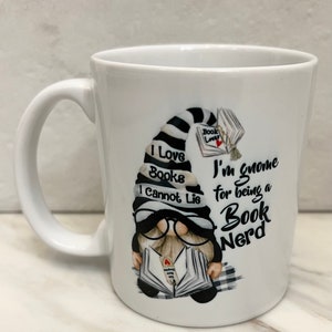 May include: White ceramic mug with a gnome design. The gnome has a black and white striped hat and glasses, holding a book. Text on the mug reads "I Love Books I Cannot Lie" and "I'm gnome for being a Book Nerd."