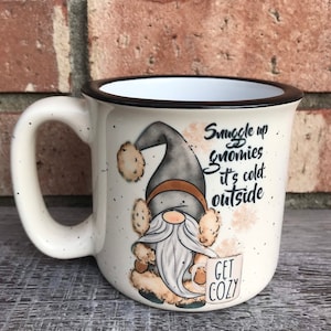 May include: White ceramic mug with a black rim and a gnome illustration. The mug has black speckles and says "Snuggle up gnomies it's cold outside" and "Get Cozy".