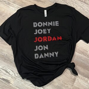 May include: Black t-shirt featuring the names DONNIE, JOEY, JORDAN, JON, and DANNY. The names are in silver glitter, with JORDAN in red glitter. The t-shirt is knotted at the hem.