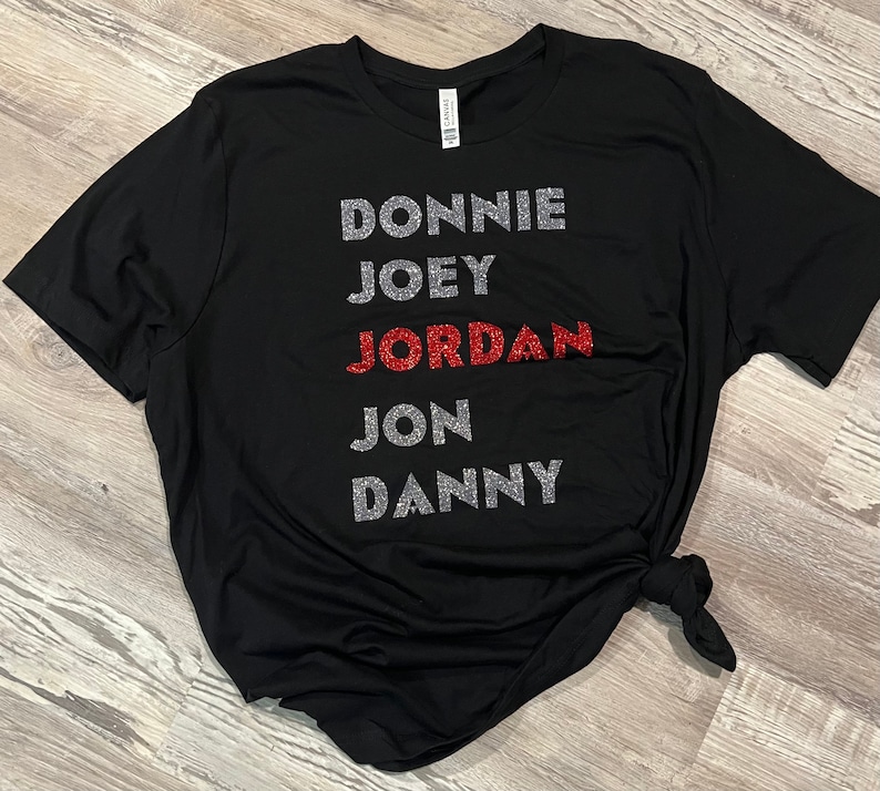 May include: Black t-shirt featuring the names DONNIE, JOEY, JORDAN, JON, and DANNY. The names are in silver glitter, with JORDAN in red glitter. The t-shirt is knotted at the hem.