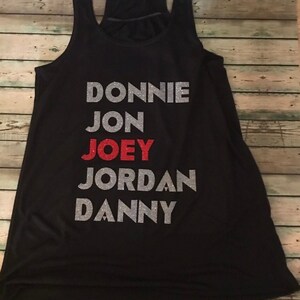 May include: Black racerback vest with the names "DONNIE", "JON", "JOEY", "JORDAN", and "DANNY" printed on the front. The names are in silver glitter, with "JOEY" in red glitter. The vest is displayed on a wooden surface.