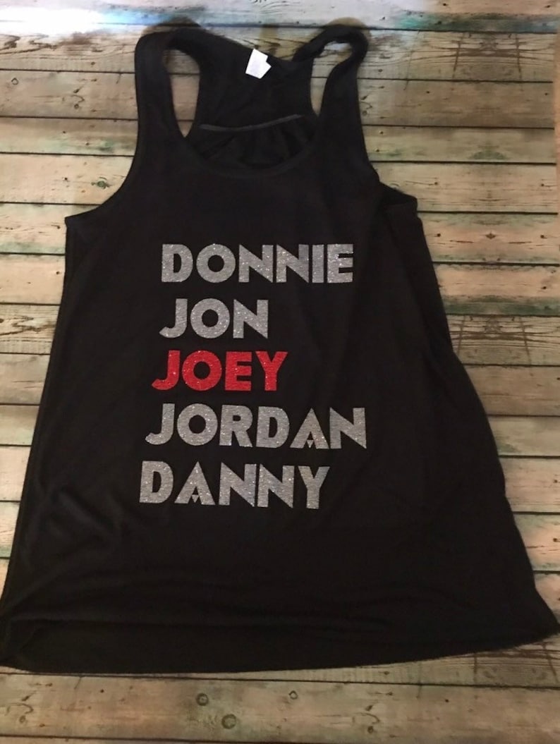 May include: Black racerback vest with the names "DONNIE", "JON", "JOEY", "JORDAN", and "DANNY" printed on the front. The names are in silver glitter, with "JOEY" in red glitter. The vest is displayed on a wooden surface.