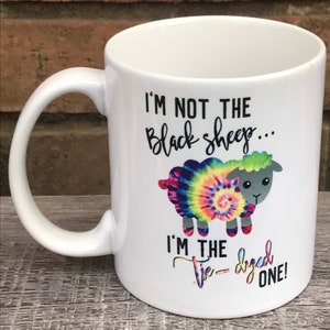May include: White ceramic mug with a black and white graphic of a tie-dye sheep. The text reads "I'm not the black sheep... I'm the tie-dyed one!"