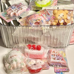 May include: A clear plastic container filled with various packaged, decorative soaps. The soaps are shaped like a bear, donuts, cake, and other desserts. The soaps are in pastel colours with decorative sprinkles and toppings.