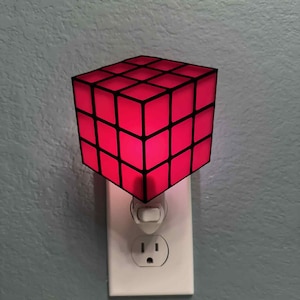 May include: A red cube-shaped night light with black grid lines is plugged into a white electrical outlet. The night light emits a soft red glow, creating a warm ambiance. The outlet is mounted on a blue-gray wall.