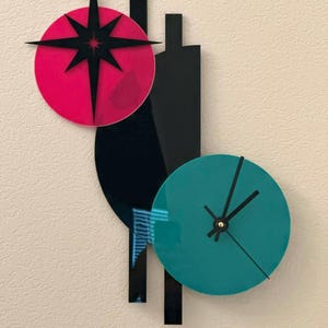 May include: A modern wall clock with a teal and black circular clock face, black hands, and a black starburst design on a red circle. The clock has a unique, abstract design with black geometric shapes.