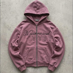Può includere: Felpa con cappuccio e zip color malva con cappuccio e tasche frontali. La parola "Mastermind" è ricamata sul petto in corsivo, con disegni di teschi e ossa incrociate sottostanti. La felpa ha una cerniera argentata.