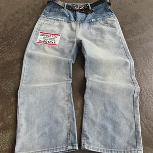 May include: Light blue denim jeans with a high waist and wide legs. The jeans have a black studded belt and a patch that reads "DOUBLE YOU TENTH SERIES Xx0010Xx". The jeans are laid flat on a gray surface.