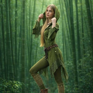 Puede incluir: Una persona con una túnica con capucha y leggings verdes está de pie sobre una roca cubierta de musgo en un bosque de bambú. La túnica tiene mangas largas y una falda fluida, combinada con botas marrones. Los leggings tienen un patrón decorativo.