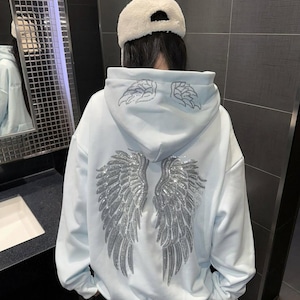 Può includere: Una felpa con cappuccio azzurra con un grande design di ali d'angelo in strass argentati sul retro. La felpa con cappuccio presenta anche disegni di ali più piccoli sul cappuccio. La persona indossa un cappello bianco e pantaloni bianchi.