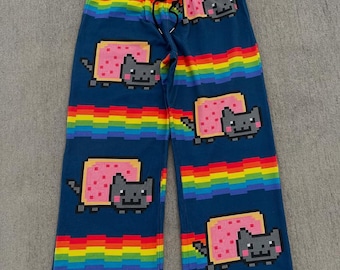 May include: Blue pajama pants with a rainbow design and pixelated Nyan Cat graphics. The pants feature a drawstring waist and multiple instances of the cat and rainbow pattern. The design includes a pink pop-tart and a gray cat.