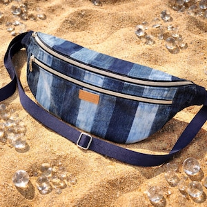 Peut inclure: Sac banane en jean bleu avec un motif rayé. Le sac comporte deux compartiments zippés avec une bordure beige et une bandoulière réglable bleu marine. Un petit patch rectangulaire marron est cousu sur le devant.