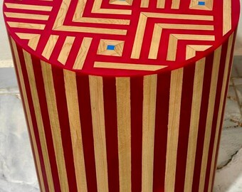 Op de afbeelding: Cilindrische houten kruk met een rood en natuurlijk houten ontwerp. De bovenkant heeft een geometrisch patroon met blauwe accenten, terwijl de zijkanten verticale rode en natuurlijke houten strepen hebben. Dit decoratieve stuk is geschikt voor diverse interieurs.