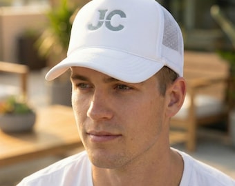 May include: White trucker hat with a curved brim and mesh back. The hat features the initials "JC" in a light blue color on the front. The hat is worn by a person in a white t-shirt.