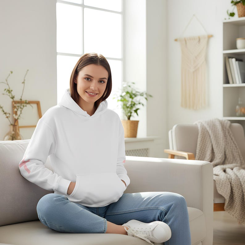 May include: A white hooded sweatshirt with a kangaroo pocket and pink accents on the sleeves. The sweatshirt is worn with blue jeans and white sneakers. The person is sitting on a gray couch in a well-lit room.
