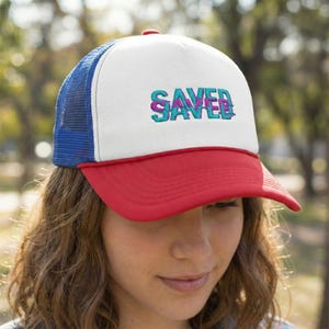 May include: A red, white, and blue trucker hat with the word "SAVED" embroidered in teal and purple. The hat features a white front panel, a red peak, and a blue mesh back.