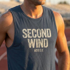 May include: A navy blue vest top featuring the text "SECOND WIND" in distressed white lettering. Below the text is "ACTS 2:2". The sleeveless shirt is made of a soft material.