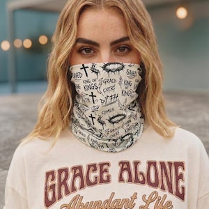 May include: Cream-colored neck gaiter with black text and cross graphics. The text includes phrases like "Saved," "King of Kings," and "Grace Every." The person is wearing a cream-colored t-shirt with the words "Grace Alone Abundant Life."