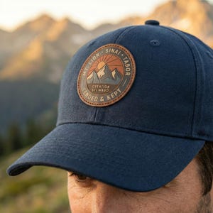 May include: Navy blue baseball cap with a circular patch featuring a mountain scene, sun, and text that reads "MT. LION SINAI + TABOR CREATION STEWARD TENDED & KEPT". The cap has a curved brim and a button on top.