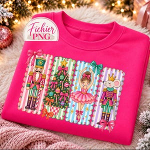 May include: A bright pink sweatshirt featuring a graphic print of four vertical panels. The panels display a nutcracker, a decorated Christmas tree, a ballerina in a pink tutu, and a mouse soldier, all accented with bows and stars.