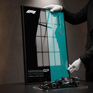 May include: A framed rectangular poster featuring the F1 logo, the Mercedes-AMG Petronas logo, and the text Mercedes GP Petronas Formula One Team. A miniature black and teal Formula One race car model sits on a clear base in front of the poster.