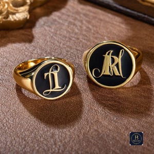 Op de afbeelding: Twee goudkleurige zegelringen met zwarte emaille gezichten. Eén ring heeft de letter "L" in een cursief lettertype en de andere toont de letters "AR". De ringen liggen op een bruin oppervlak.